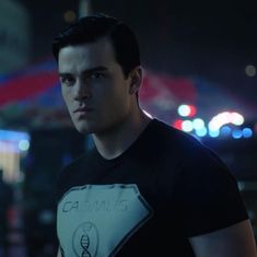 a man in a black t - shirt is staring at the camera with city lights behind him