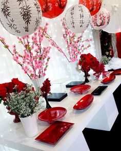 a table with red plates and vases filled with flowers on top of it, surrounded by hanging lanterns