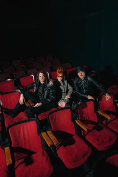 three men sitting on red chairs in an empty theater with their arms around each other