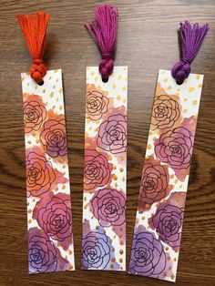 three bookmarks decorated with flowers and tassels on top of a wooden table