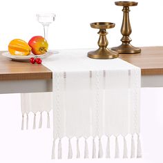 a white table runner with tassels and fruit on it next to two candles