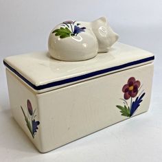 a white ceramic box with flowers painted on the side and a cat figurine sitting on top