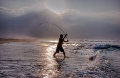 a man standing on top of a beach next to the ocean holding a fishing pole