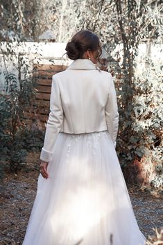 a woman in a white dress and jacket walking through the woods with her back to the camera