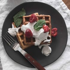 a waffle topped with whipped cream and raspberries on a black plate next to a fork