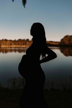 a pregnant woman standing in front of a body of water at sunset with her back to the camera