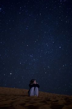 a man sitting on top of a sandy beach under a night sky filled with stars