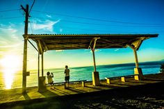 two people standing under a covered area next to the ocean on a sunny day at sunset