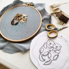 two otters are hugging each other in this embroidery project with scissors and thread on the table