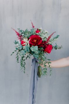 a woman holding a bouquet with red flowers and greenery in her hand, against a gray wall