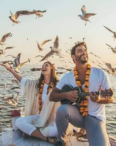 a man and woman sitting on a boat with seagulls flying above them while playing the guitar