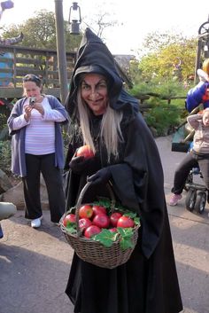 a woman dressed as a witch holding a basket of apples and vegetables in her hands