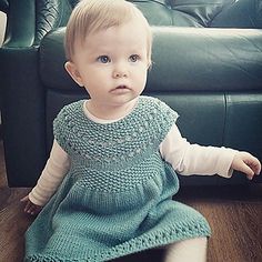 a baby sitting on the floor wearing a blue knitted dress and white socks, looking at the camera