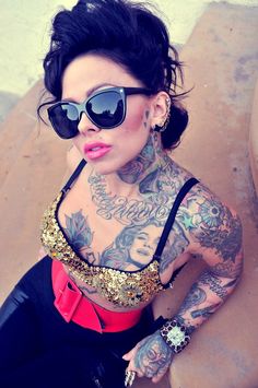 a woman with tattoos on her body wearing black sunglasses and a red top is sitting down