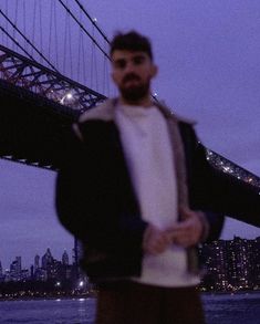 a man standing in front of a bridge with the city lights behind him at night