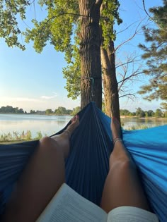 a person laying in a hammock reading a book next to a tree and water