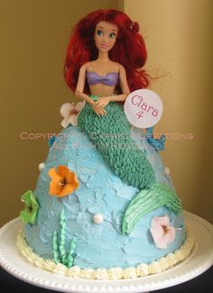 there is a cake that looks like a mermaid sitting on top of a rock and holding a name plate