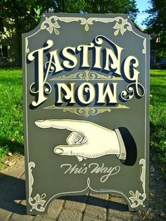 a sign that says tasting now in front of some green grass and trees with a hand pointing at the viewer