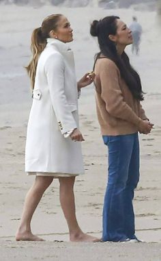 two women standing on the beach talking to each other and one is wearing a white coat