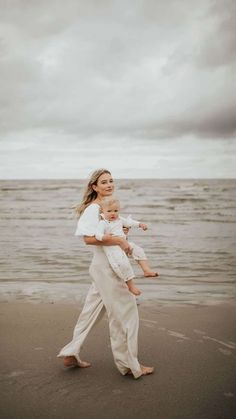 a woman holding a baby walking on the beach in front of the ocean under a cloudy sky