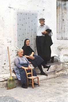 three people sitting on the steps in front of an old building, one woman is holding a broom