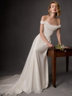 a woman sitting on top of a wooden table wearing a white dress and holding flowers