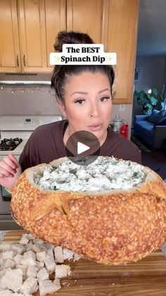 a woman is holding a large bread bowl with spinach dip in it and looking at the camera