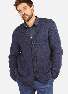 a man wearing a blue cardigan sweater standing in front of a white wall with his hands in his pockets