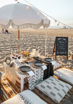 a table set up on the beach for an outdoor party with food and drinks under an umbrella