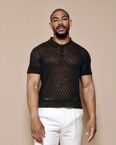 a man standing in front of a beige wall wearing white pants and a black shirt