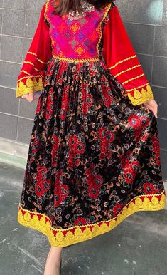 This vibrant traditional dress showcases a bold pink handmade top with colorful square patterns and long red sleeves edged with golden details. The black skirt is patterned with red and grey flowers, highlighted by a bright yellow hemline featuring gold floral designs. This dress combines simplicity with elegance, making it perfect for cultural events or as a unique addition to any collection for those who appreciate a touch of tradition. Red Dresses With Traditional Patterns For Festivals, Red Bohemian Dress For Transition Seasons, Ceremonial Red Dress With Traditional Patterns, Bohemian Red Dress For Seasonal Wear, Red Dresses With Embroidered Border For Traditional Ceremonies, Red Bohemian Ceremonial Dress, Red Bohemian Dress For Ceremonial Occasions, Red Long Sleeve Dress With Traditional Patterns, Red Dress With Dabka For Traditional Ceremonies