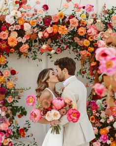 a bride and groom kissing in front of a floral arch with pink, orange and yellow flowers