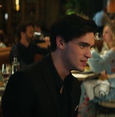 a man sitting at a table in a restaurant looking off into the distance with other people behind him