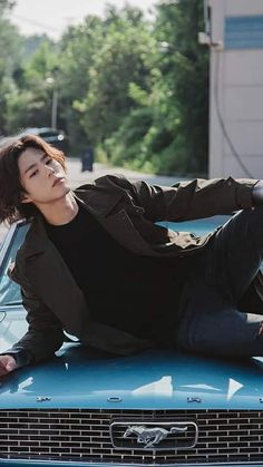 a young man sitting on the hood of a blue car with trees in the background