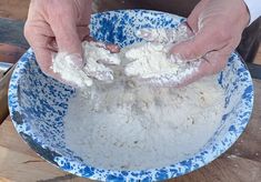 two hands are kneading flour in a blue bowl on a wooden table top