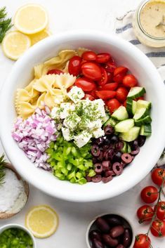 a white bowl filled with pasta salad next to olives, tomatoes and cucumbers