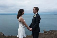 a man and woman standing next to each other on top of a hill near the ocean