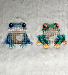 two cross stitch frogs sitting next to each other on a white surface with one frog looking at the camera