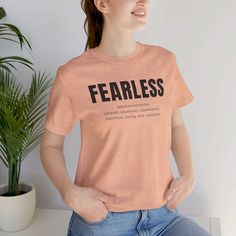 a woman sitting on a table with her hands in her pockets and wearing a peach shirt that says fearless