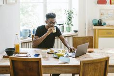 a man sitting at a table with a laptop eating food and drinking from a cup