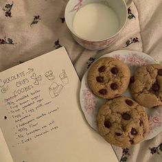 three chocolate chip cookies on a plate next to a cup of milk and an open book