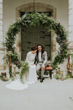 a bride and groom sitting on a bench in front of an arch with greenery