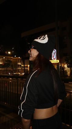 a woman wearing a black hat standing next to a fence at night with buildings in the background