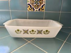 a white dish with green designs on it sitting on a blue tile wall next to a mirror