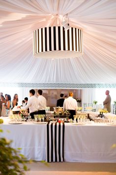 people standing around a table with food and wine glasses on it, in front of a white tent