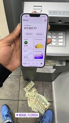 a person holding up an iphone in front of a atm machine with cash on the floor