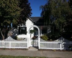 a white picket fence in front of a house with trees and bushes on the side