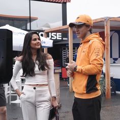 a man standing next to a woman in white pants and an orange jacket on the street