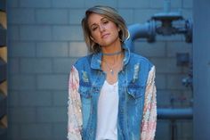 a woman standing in front of a brick wall wearing a jean jacket and white top