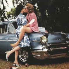 two women are sitting on the hood of an old car and kissing each other with their legs crossed